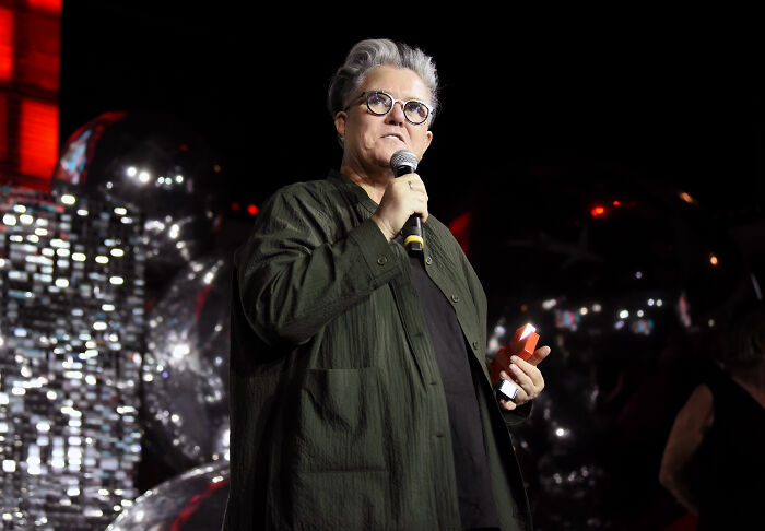 Celebrity speaking at event, holding microphone and notes, related to celebrities who slammed Donald Trump in 2025.
