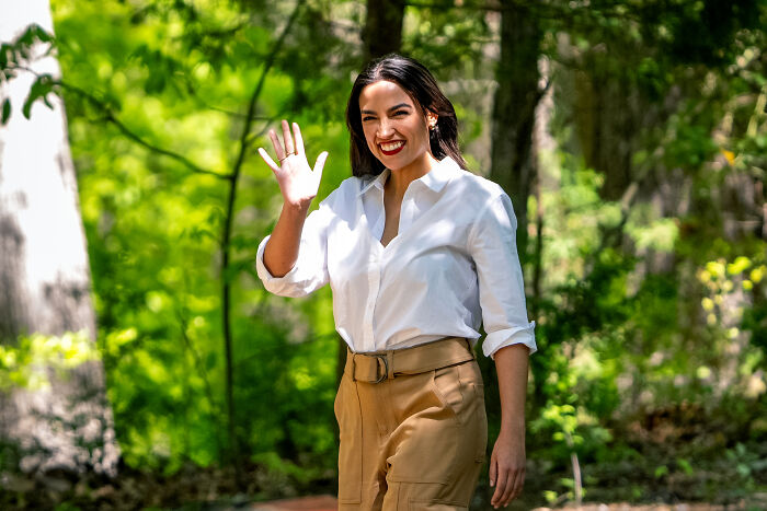 AOC smiling and waving outdoors, linked to reports of spending on luxury hotels, fine dining, and Puerto Rico venue expenses.