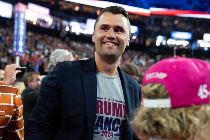 Charlie Kirk smiling at a political rally, wearing a Trump 2024 shirt amid a crowd of supporters.