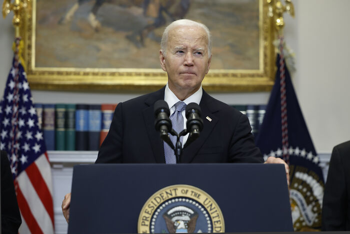 Joe Biden standing at a podium with presidential seal, speaking in front of American flags and bookshelves.