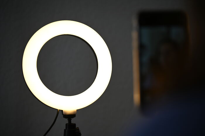 Ring light glowing brightly on a stand with a blurred person holding a smartphone nearby for recording or streaming.