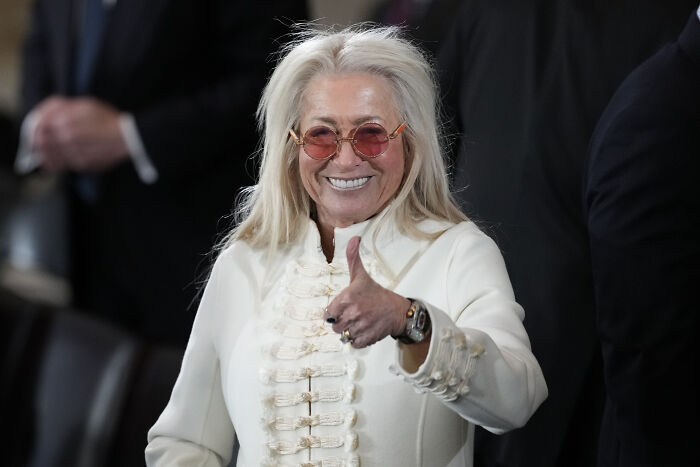 Woman in white jacket and orange glasses giving thumbs up, illustrating billionaire donor offering Trump $250 million.