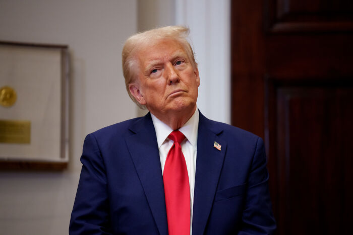 Former President Trump in a dark suit and red tie, looking serious during an indoor event with wood panel background.