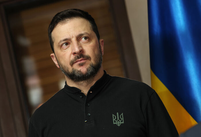 Man in black shirt with Ukrainian emblem standing near Ukrainian flag, symbolizing explosive political scandals in 2025.