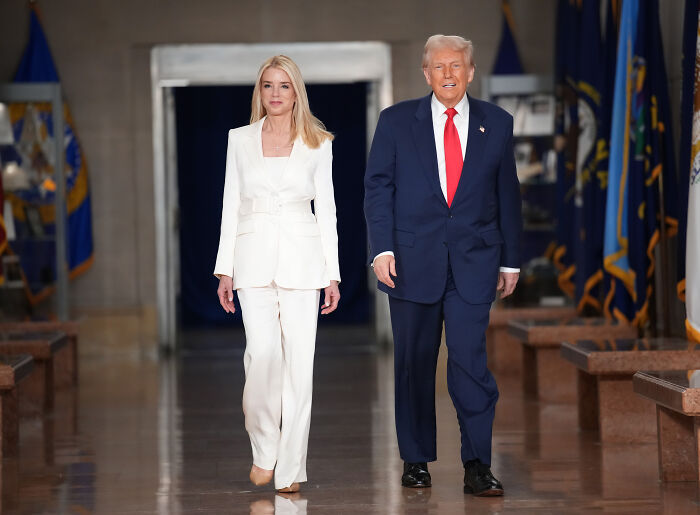 Pam Bondi walking alongside a man in a suit in a hallway with flags, appearing confident and formal.