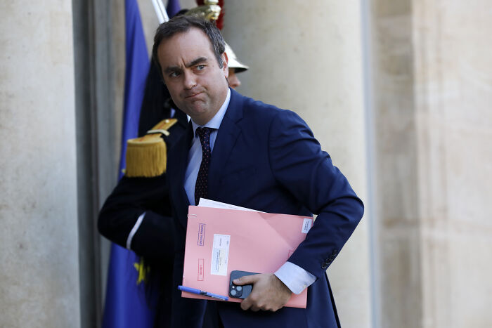 Man in a navy suit holding pink folders and a phone, leaving a building related to political scandals in 2025.