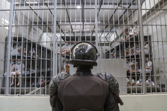 A guard in riot gear faces a large group of detainees behind bars highlighting issues of corporate censorship and deportation.