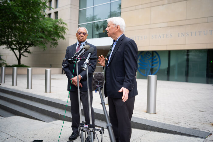 Two men speaking at a press conference outside the U.S. Institute of Peace with microphones and cameras present.