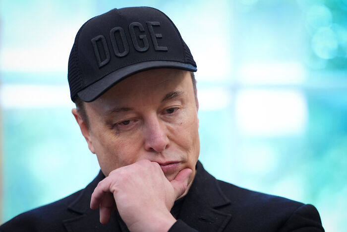 Person wearing a black DOGE cap and black jacket, appearing thoughtful with hand on chin against a blurred background.