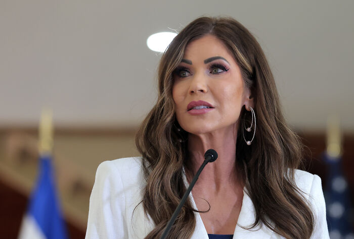 Kristi Noem speaking at a podium with a microphone, wearing hoop earrings and a white blazer indoors.