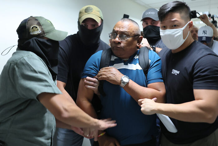 A man in glasses being escorted by masked men in a tense scene related to political scandals of 2025.