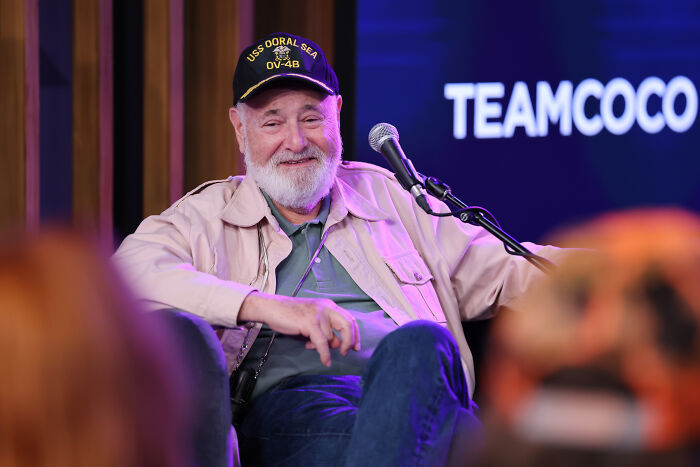Rob Reiner wearing a cap and jacket, speaking into a microphone during a live event discussing Charlie Kirk assassination.