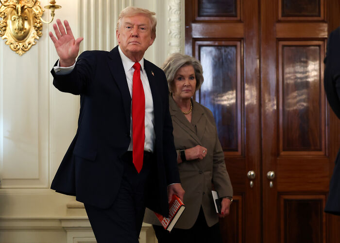 Donald Trump in a suit waving, with his chief of staff holding a notebook, inside a formal room with wooden doors.
