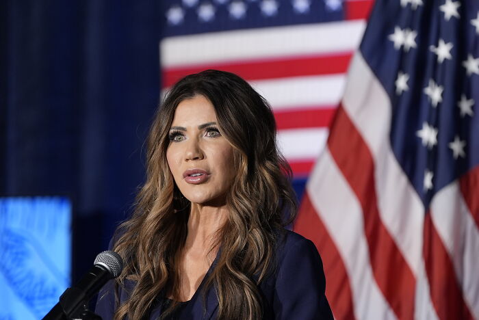 Kristi Noem speaking at event with American flags in background during hot mic moment involving TSA staff fear.