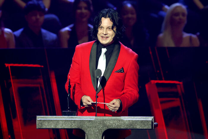 Musician in red jacket speaking at a podium with audience blurred in the background, related to celebrities slamming Donald Trump.