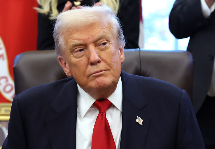 Donald Trump sitting in an office wearing a dark suit and red tie during a formal event with an American flag pin.
