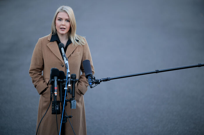 Karoline Leavitt standing outdoors in a brown coat, speaking into multiple microphones during a public statement.