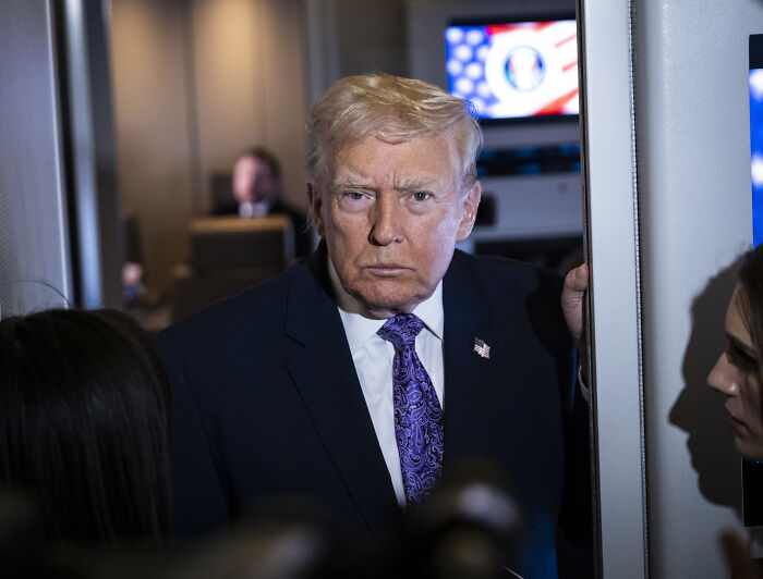 Trump in a dark suit and purple tie, appearing serious while interacting with reporters during an indoor event.