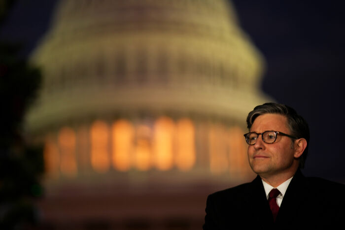 House Speaker Mike Johnson in a dark suit and glasses standing outside the Capitol building at night, focused and resolute.