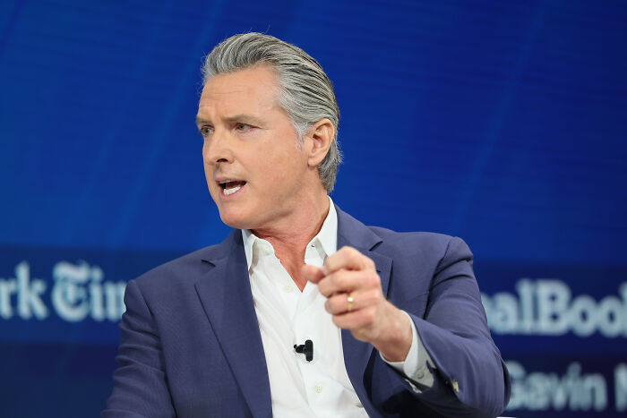 Gavin Newsom speaking at an event, wearing a navy suit and white shirt, with a serious expression on stage.
