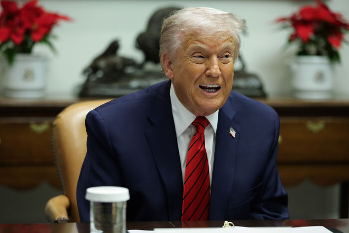 Former President Trump sitting at a table indoors, smiling and wearing a navy suit with red striped tie.
