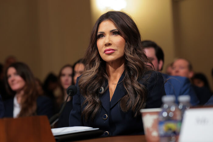Kristi Noem testifying at a hearing, dressed in dark blazer, with focused expression and audience in background.