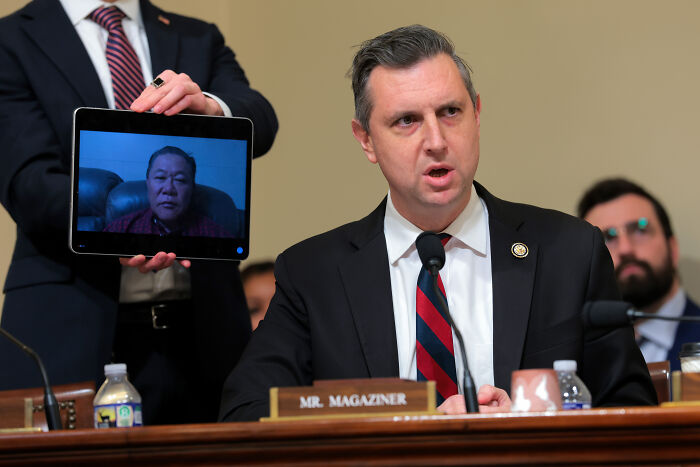 Congressman speaking at a hearing with a tablet showing a deported veteran joining remotely during veteran deportation testimony.