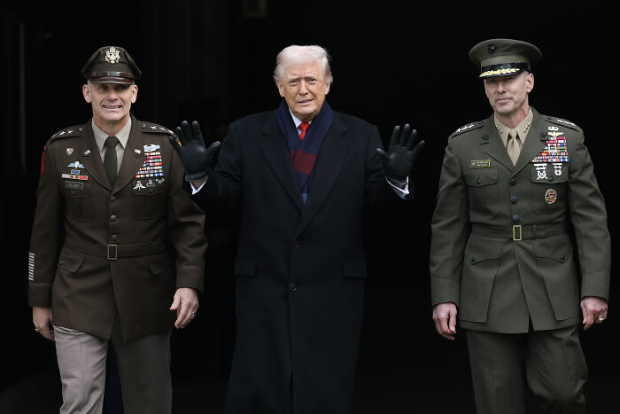 Donald Trump standing between two military officers, wearing a dark coat and gloves, related to Trump&rsquo;s war crimes in the Caribbean.