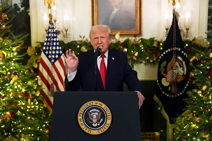 Donald Trump speaking on prime-time TV, standing at presidential podium with flags and Christmas decorations.