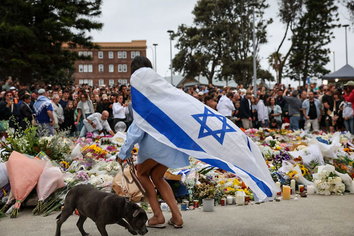 Woman draped in Israeli flag at Bondi Beach memorial with crowd honoring victims after Hanukkah attack gunman incident.