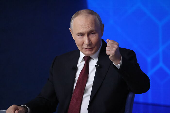 Vladimir Putin in a dark suit and maroon tie making a fist gesture during a formal discussion against a blue background.