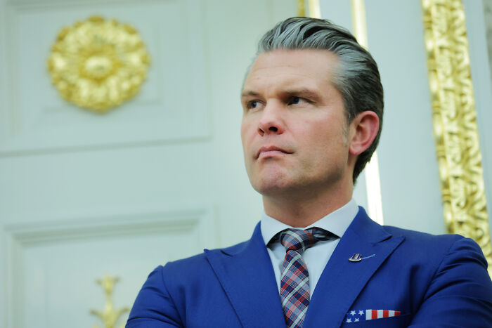 Man in a blue suit with an American flag pocket square, standing in a formal room with gold accents and decorations