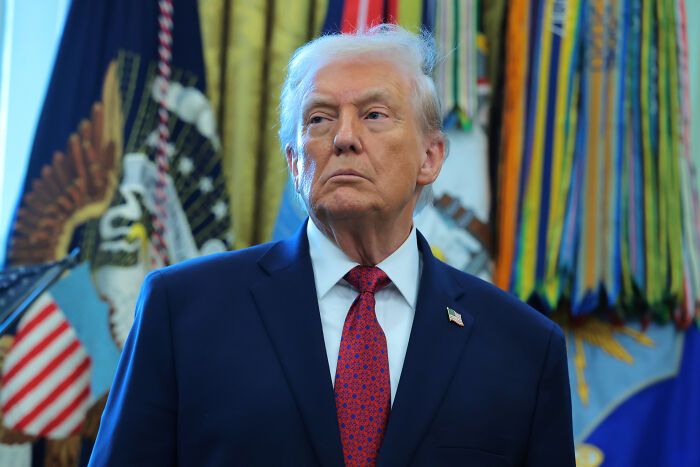 Donald Trump in a navy suit and red tie, standing in the Oval Office with flags behind, amid lawmakers warning Pam Bondi prosecution.