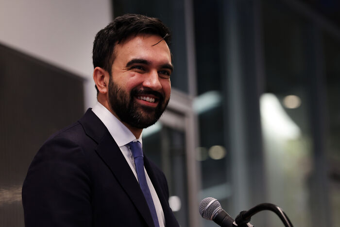 Smiling man in a suit speaking at a microphone during an indoor event related to Kim Petras and Zohran Mamdani.
