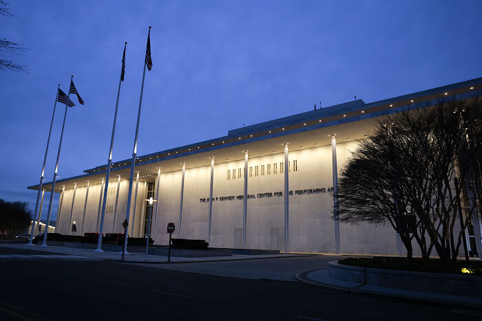 Exterior view of the Kennedy Center at dusk with flags and illuminated facade referencing Trump rename vote controversy.