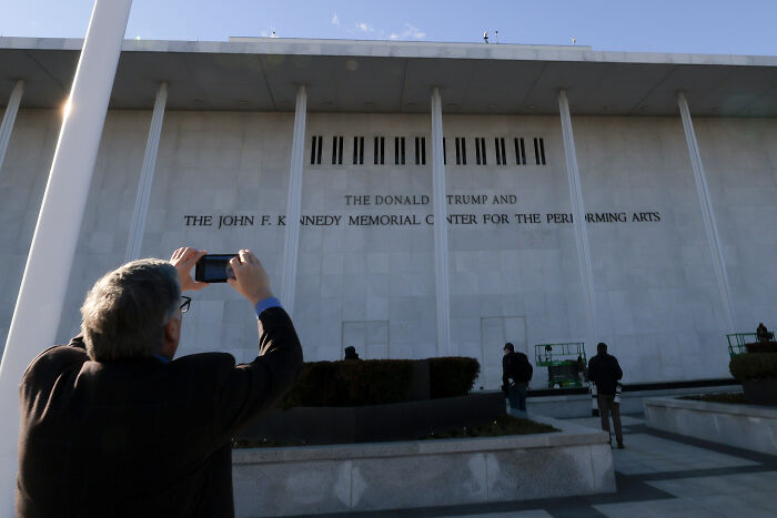 Man taking photo of Kennedy Center building facade showing Trump rename signage amid New Year&rsquo;s cancellation and backlash.