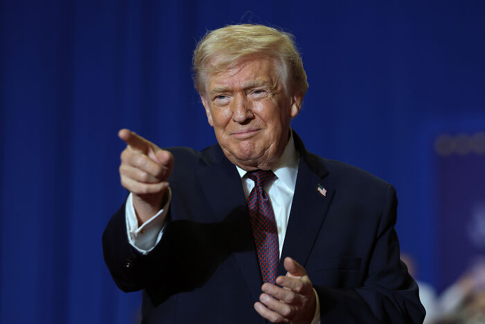 Donald Trump in a dark suit pointing forward during a public speaking event related to Nicki Minaj and JD Vance discussion.