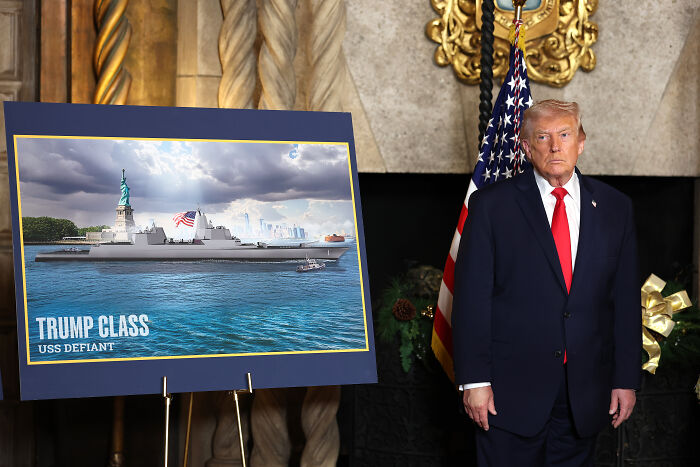 Former President Trump standing next to a display of a new battleship named Trump Class USS Defiant in a formal room.