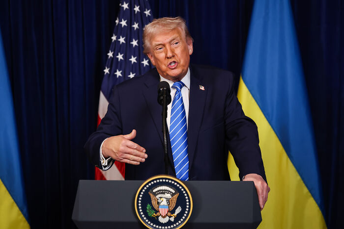 Former President Trump speaking at a podium with US and Ukraine flags, Zelenskyy&rsquo;s face reaction implied in context.