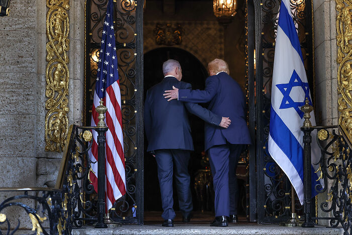 Two men walking into ornate building entrance with US and Israeli flags, illustrating Iran warns severe response after strike threats.