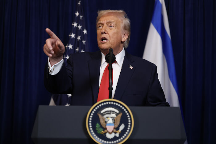 Donald Trump at podium speaking with American and Israeli flags in background, related to Jack White AI video controversy.