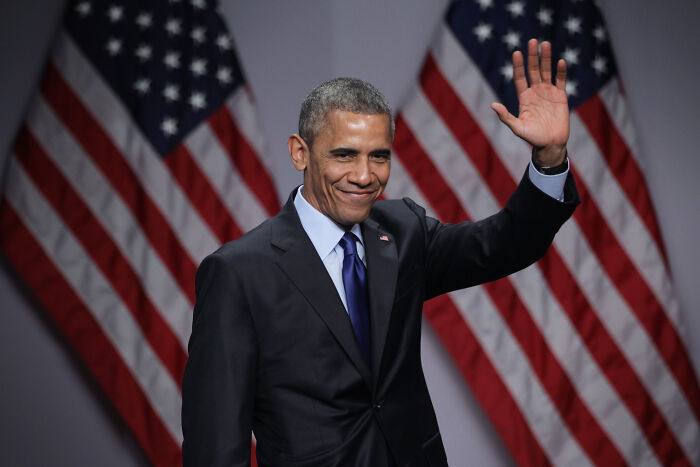 Barack Obama waving with U.S. flags in the background during a formal event, wearing a dark suit and blue tie.