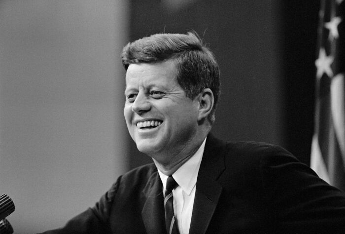 Black and white photo of JFK smiling and speaking at a podium with the American flag in the background.