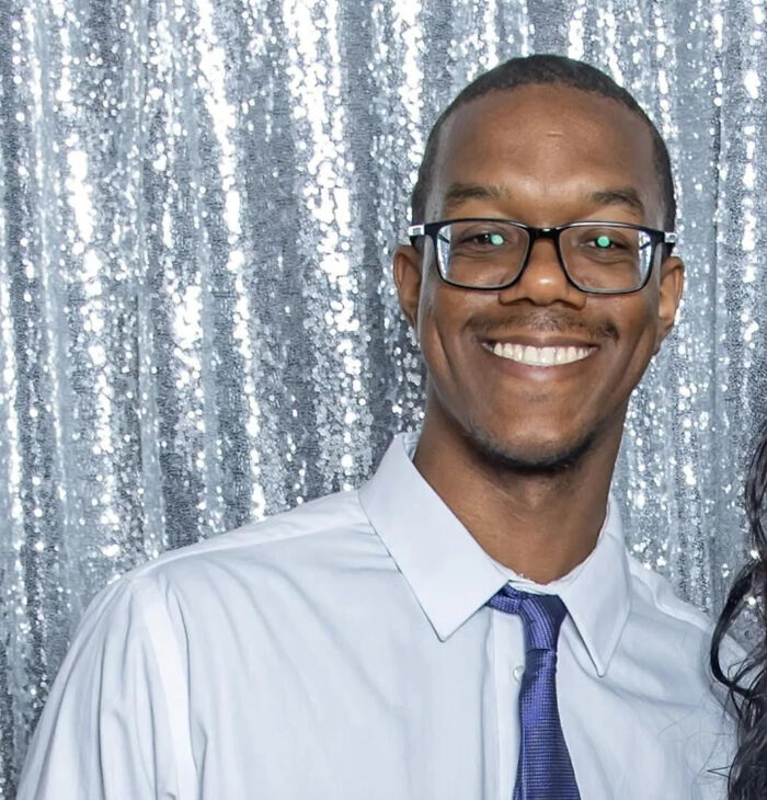 Smiling man wearing glasses, a white shirt, and blue tie standing in front of a silver sequin backdrop at an event.