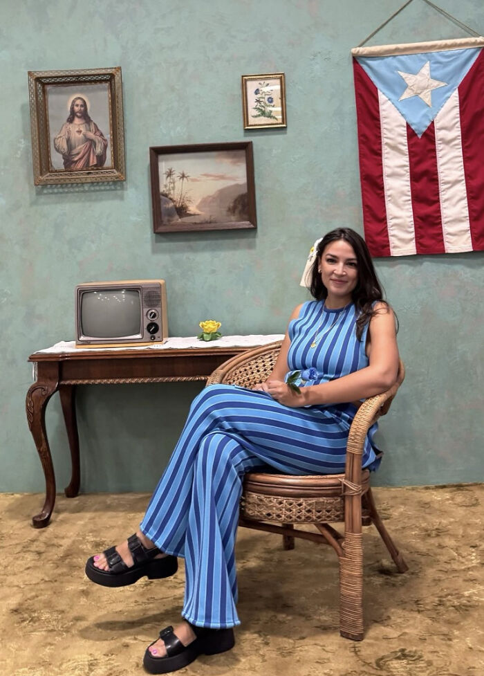 AOC seated indoors near Puerto Rico flag, vintage TV, and paintings, relating to luxury hotels and Puerto Rico venue visit.