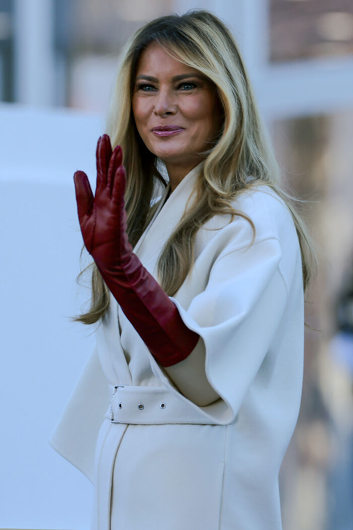 Melania Trump wearing a white coat and red gloves, smiling and waving outdoors in a candid moment.