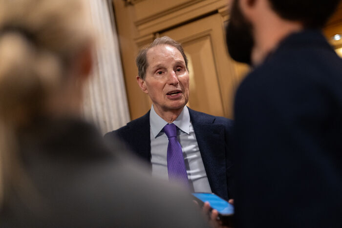 Man in a suit and purple tie speaking to two people in a hallway, related to Epstein bank records and island photos discussion.