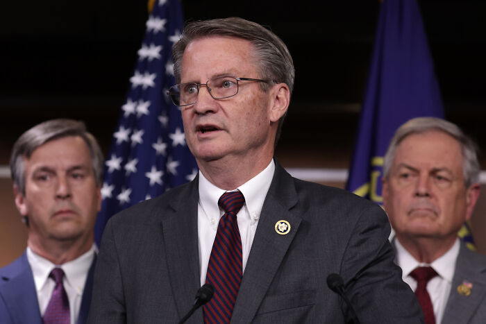 A man in a suit and glasses speaking at a podium with American and dark blue flags behind him, discussing Taliban funding.
