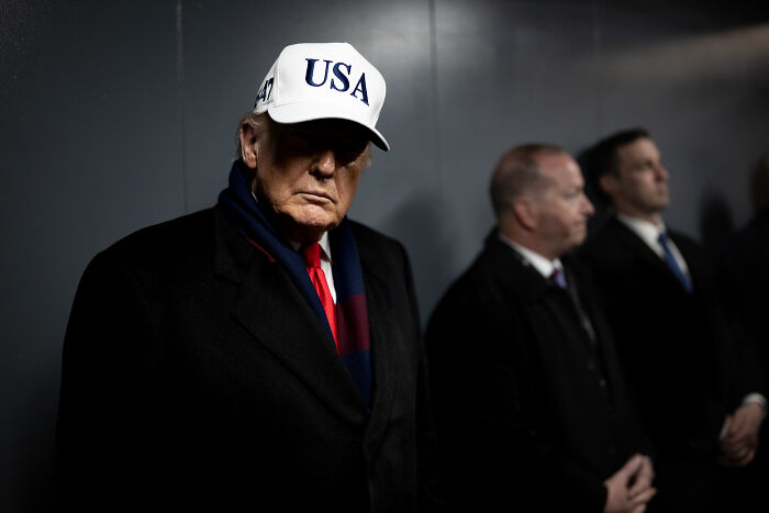 Former president wearing USA hat and dark coat, with two men blurred in background during a staged TV address setting.