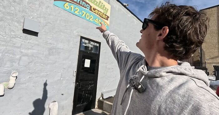 Man in a gray hoodie pointing at a Minnesota child care center sign, highlighting child care funds and fraud issues.